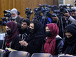 Mehrere Journalist*innen stehen und sitzen in einer Presekonferenz. Die sitzenden Frauen tragen Kopftücher sowie medizinische Masken, die dahinter sitzenden und stehenden Männer allesamt nicht.